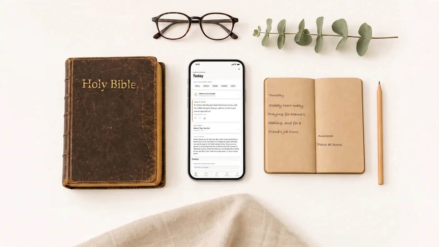A worn leather Bible lettered Holy Bible, reading glasses, a eucalyptus sprig, an open notebook with a handwritten prayer note, a pencil, and a phone showing the app's Today screen, laid out on a pale table.